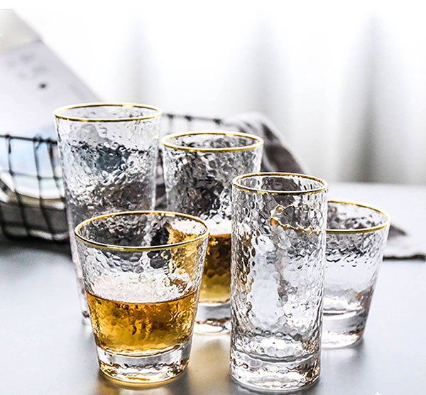 C&aacute;c ly thủy tinh trong suốt với viền v&agrave;ng, một số chứa chất lỏng m&agrave;u hổ ph&aacute;ch, được đặt tr&ecirc;n b&agrave;n.