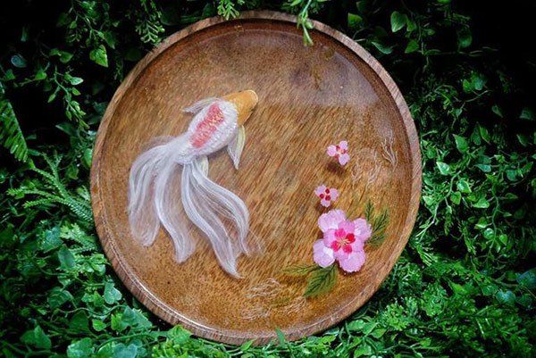 Đĩa tròn gỗ có hình cá vàng và hoa hồng trang trí, đặt trên nền lá xanh tươi.