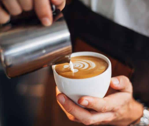 Người đang r&oacute;t sữa tạo h&igrave;nh nghệ thuật v&agrave;o t&aacute;ch c&agrave; ph&ecirc;, thể hiện kỹ thuật pha chế tinh tế.