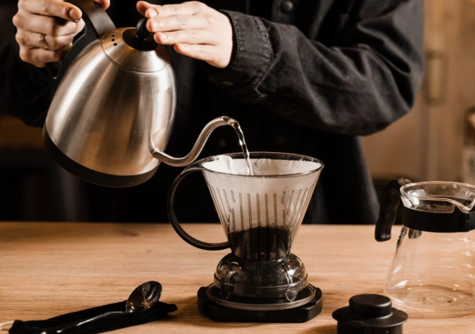 Người đang rót nước từ ấm đun vào phễu pha cà phê trên bàn gỗ, bên cạnh là bình đựng.