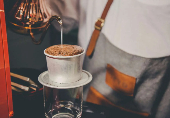 Người pha chế đang rót nước vào phin cà phê đặt trên ly thủy tinh trong suốt.