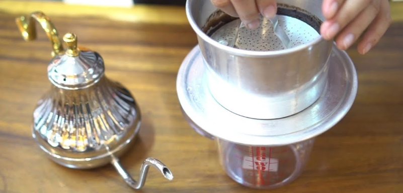 Người pha cà phê phin truyền thống trên bàn gỗ, bên cạnh là ấm nước kim loại.