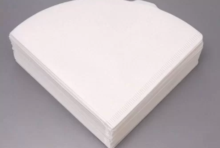 Chồng giấy lọc c&agrave; ph&ecirc; m&agrave;u trắng h&igrave;nh quạt đặt tr&ecirc;n bề mặt phẳng m&agrave;u x&aacute;m.