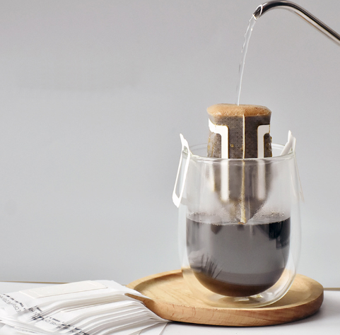 Người pha c&agrave; ph&ecirc; bằng c&aacute;ch r&oacute;t nước n&oacute;ng qua t&uacute;i lọc treo tr&ecirc;n miệng ly thủy tinh đặt tr&ecirc;n đĩa gỗ.