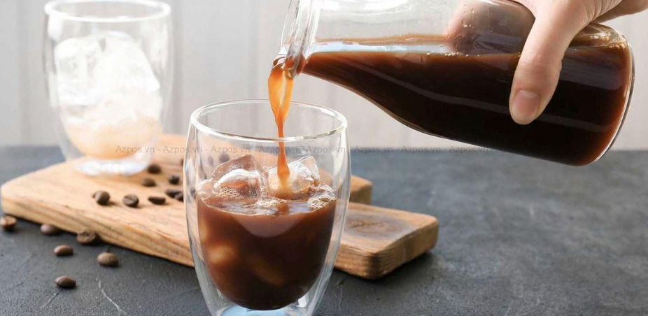 Người r&oacute;t c&agrave; ph&ecirc; v&agrave;o ly thủy tinh c&oacute; đ&aacute;, b&ecirc;n cạnh l&agrave; một ly kh&aacute;c v&agrave; v&agrave;i hạt c&agrave; ph&ecirc; tr&ecirc;n mặt b&agrave;n.