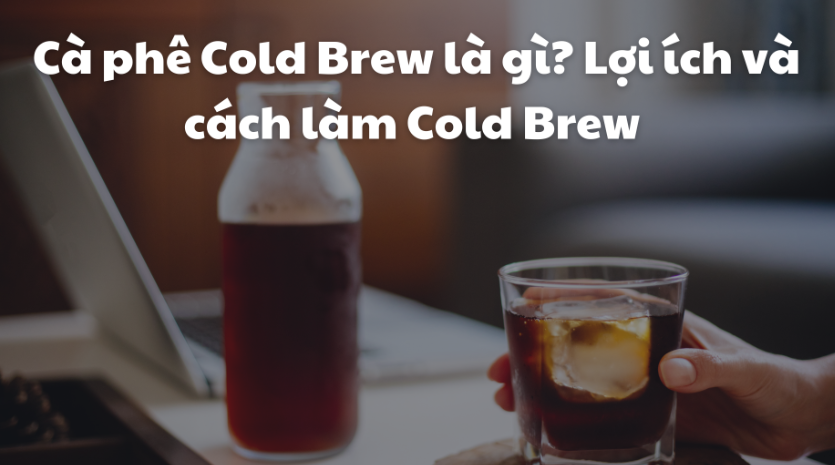 Một tay cầm ly c&agrave; ph&ecirc; cold brew với đ&aacute;, cạnh đ&oacute; l&agrave; chai cold brew v&agrave; m&aacute;y t&iacute;nh x&aacute;ch tay tr&ecirc;n b&agrave;n.