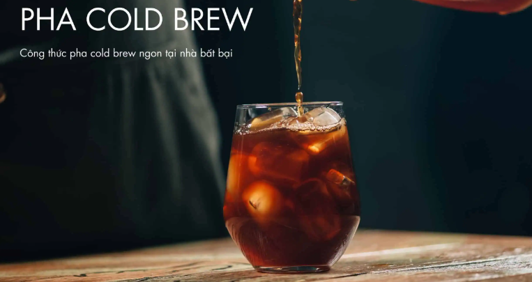 Ly cold brew đang được r&oacute;t đầy, đặt tr&ecirc;n b&agrave;n gỗ, nền tối. Văn bản: Pha Cold Brew.