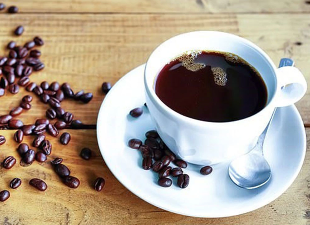 T&aacute;ch c&agrave; ph&ecirc; tr&ecirc;n đĩa trắng, cạnh l&agrave; hạt c&agrave; ph&ecirc; v&agrave; th&igrave;a bạc, đặt tr&ecirc;n b&agrave;n gỗ.