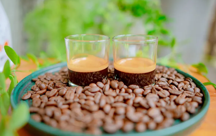 Hai ly c&agrave; ph&ecirc; espresso tr&ecirc;n đĩa đầy hạt c&agrave; ph&ecirc;, nền xanh mờ.