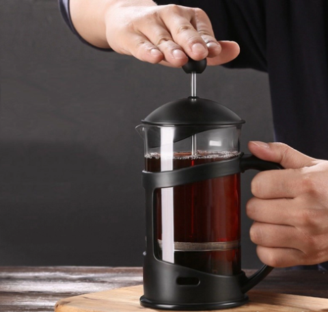 Một người đang sử dụng b&igrave;nh pha c&agrave; ph&ecirc; kiểu Ph&aacute;p để pha chế đồ uống, đặt tr&ecirc;n b&agrave;n gỗ.