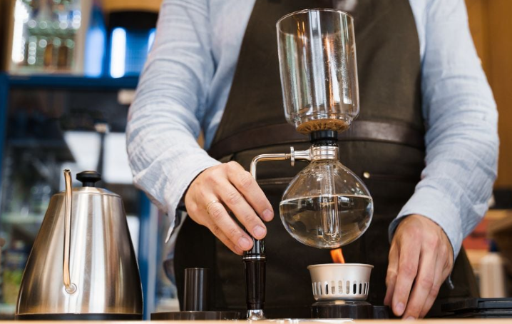 Người đang pha c&agrave; ph&ecirc; bằng dụng cụ pha chế siphon trong một qu&aacute;n c&agrave; ph&ecirc;, b&ecirc;n cạnh l&agrave; ấm đun nước kim loại.