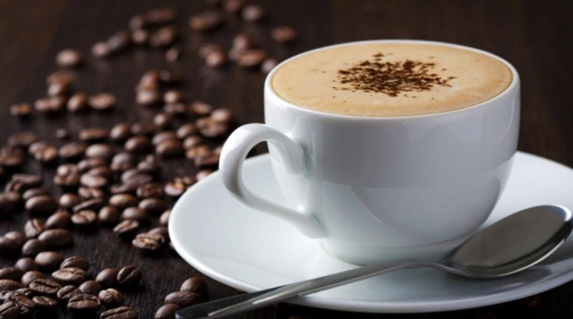 Tách cà phê cappuccino trên đĩa trắng, bên cạnh là muỗng kim loại và hạt cà phê rải rác trên bàn gỗ.