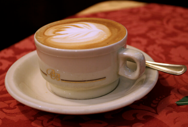 Tách cappuccino với lớp bọt sữa tạo hình nghệ thuật, đặt trên đĩa sứ trắng, nền khăn trải bàn màu đỏ.