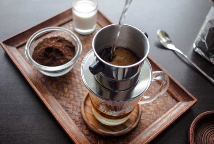 Pha c&agrave; ph&ecirc; bằng phin tr&ecirc;n khay gỗ, b&ecirc;n cạnh l&agrave; bột c&agrave; ph&ecirc;, sữa v&agrave; th&igrave;a kim loại.