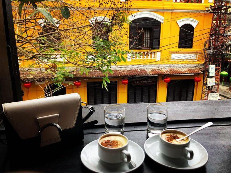 Khám Phá Gu Cà Phê Nguyên Chất - Xu Hướng Thưởng Thức Hiện Đại Của Giới Trẻ! 10 Hai tách cà phê trên bàn cạnh cửa sổ, nhìn ra ngoài là ngôi nhà màu vàng với cửa sổ đen và cây xanh.
