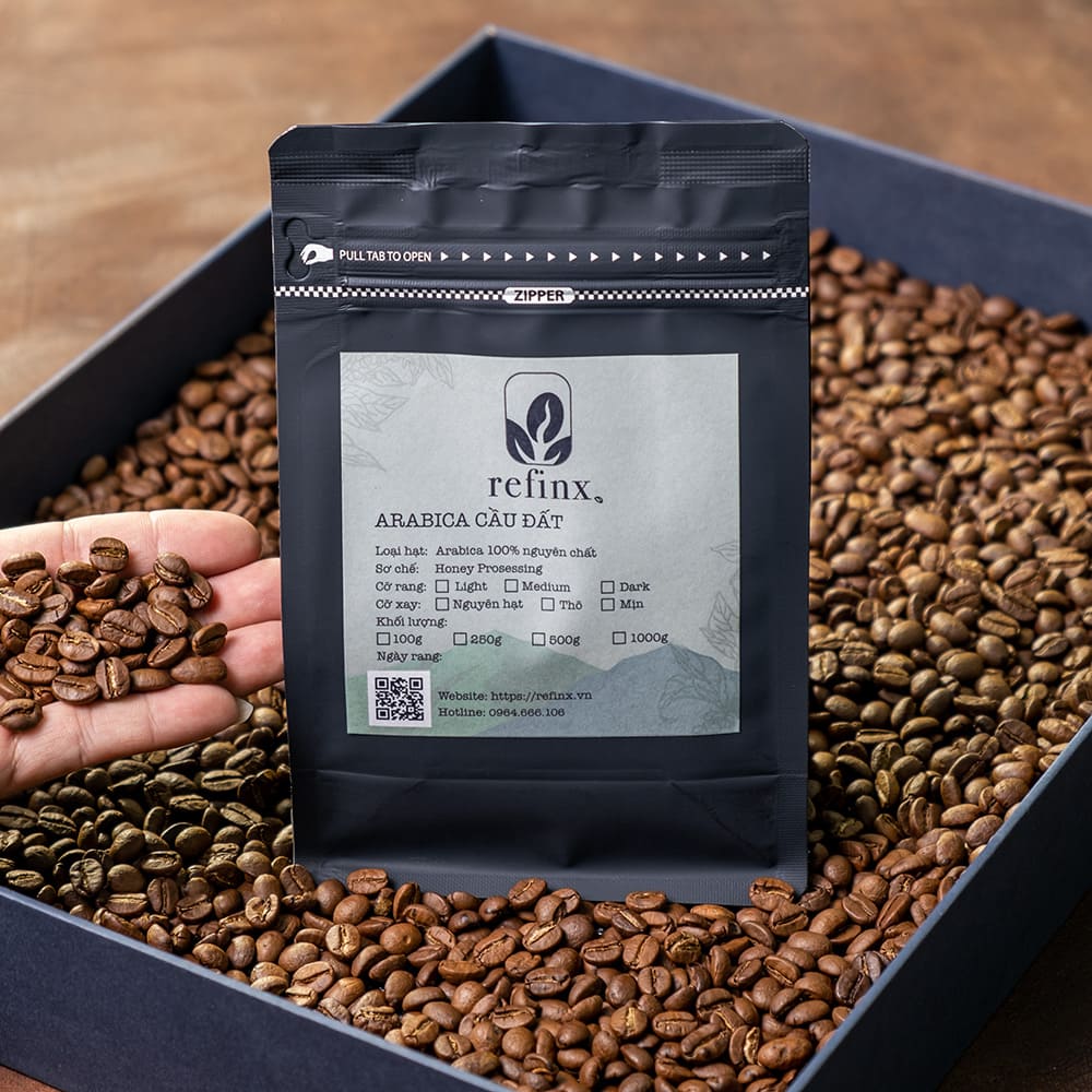 T&uacute;i c&agrave; ph&ecirc; Arabica Cầu Đất đặt tr&ecirc;n hạt c&agrave; ph&ecirc;, một tay cầm một nắm hạt c&agrave; ph&ecirc;.