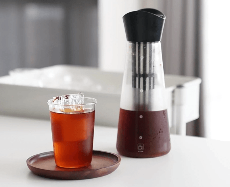 Khám Phá Thế Giới Dụng Cụ Pha Cà Phê Đỉnh Cao Cho Tín Đồ Yêu Ngâm Ủ (Phần I) 12 Cốc nước đá lạnh trên đĩa gỗ cạnh bình nước trong suốt có nắp đen, đặt trên bàn trắng.