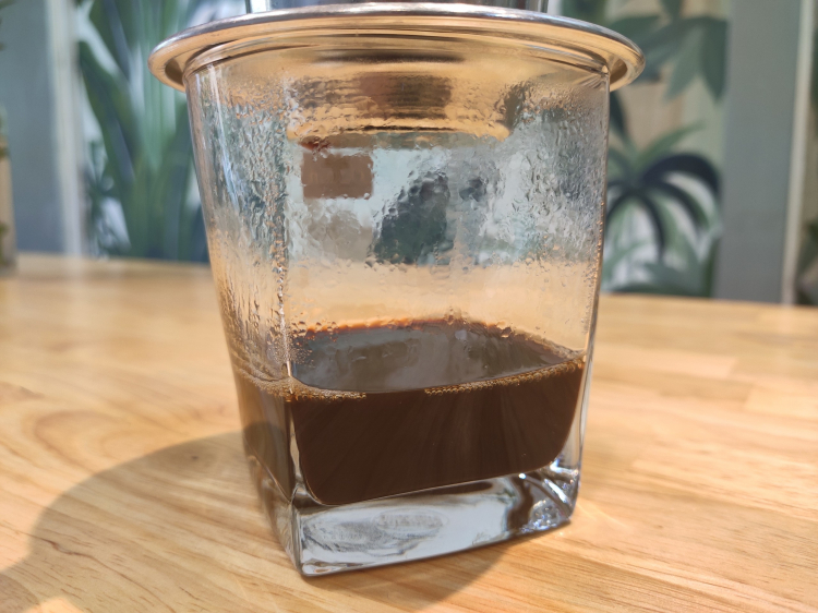 Ly cà phê phin đang nhỏ giọt, đặt trên bàn gỗ, nền có cây xanh.