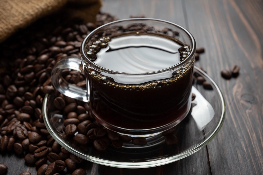 Tách cà phê đen trên đĩa thủy tinh, xung quanh là hạt cà phê, trên nền gỗ tối màu.