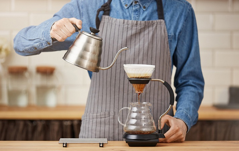Khám Phá Bí Ẩn Của Moka Pot: Hành Trình Đầy Mê Hoặc Với Bình Pha Cà Phê Huyền Thoại 3 Người mặc tạp dề đang pha cà phê bằng dụng cụ pha chế thủ công trên bàn gỗ trong không gian bếp.