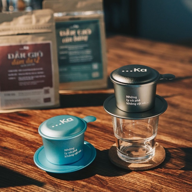 Bộ phin cà phê và ly thủy tinh trên bàn gỗ, phía sau là hai túi cà phê.