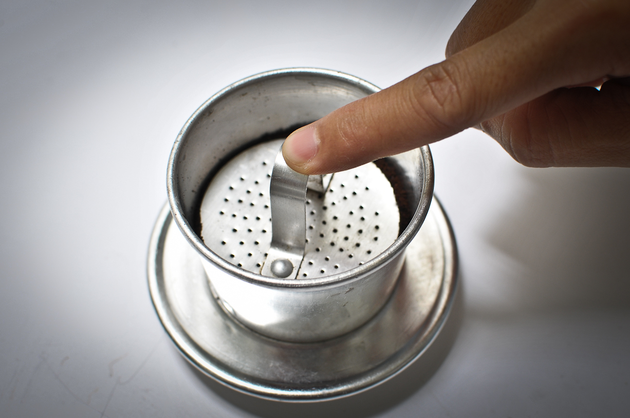 Ngón tay đặt lên nắp phin cà phê bằng kim loại trên nền trắng.