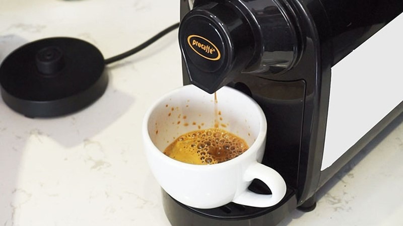 M&aacute;y pha c&agrave; ph&ecirc; đang chảy c&agrave; ph&ecirc; v&agrave;o t&aacute;ch trắng đặt tr&ecirc;n đế m&aacute;y, bề mặt t&aacute;ch c&oacute; lớp bọt n&acirc;u.