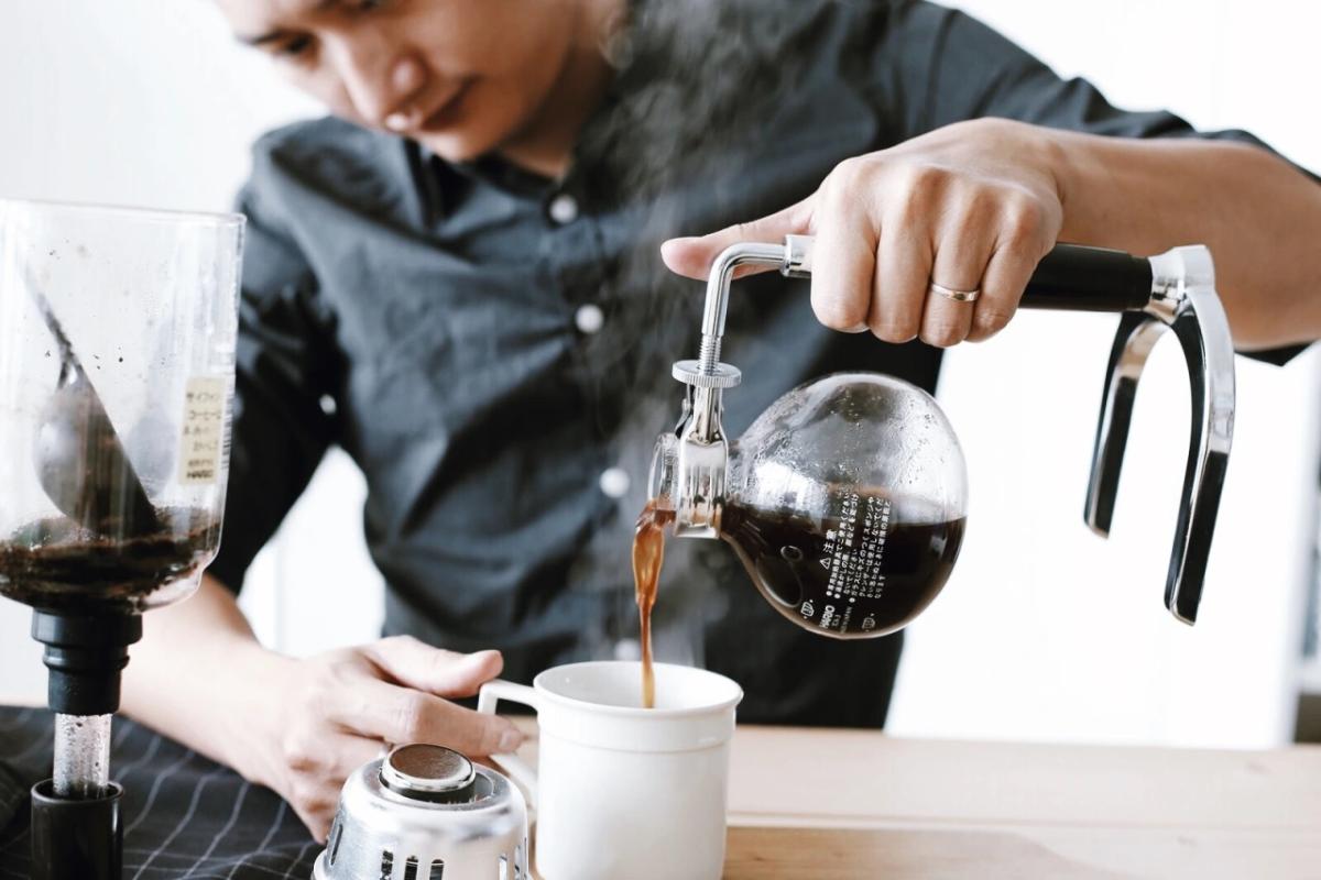 Người đ&agrave;n &ocirc;ng pha c&agrave; ph&ecirc; bằng b&igrave;nh siphon, r&oacute;t c&agrave; ph&ecirc; v&agrave;o cốc trắng tr&ecirc;n b&agrave;n gỗ.