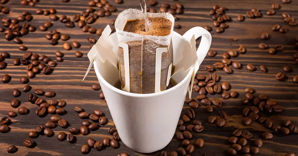 T&uacute;i lọc c&agrave; ph&ecirc; đang được pha trong cốc trắng, xung quanh l&agrave; những hạt c&agrave; ph&ecirc; tr&ecirc;n nền gỗ.