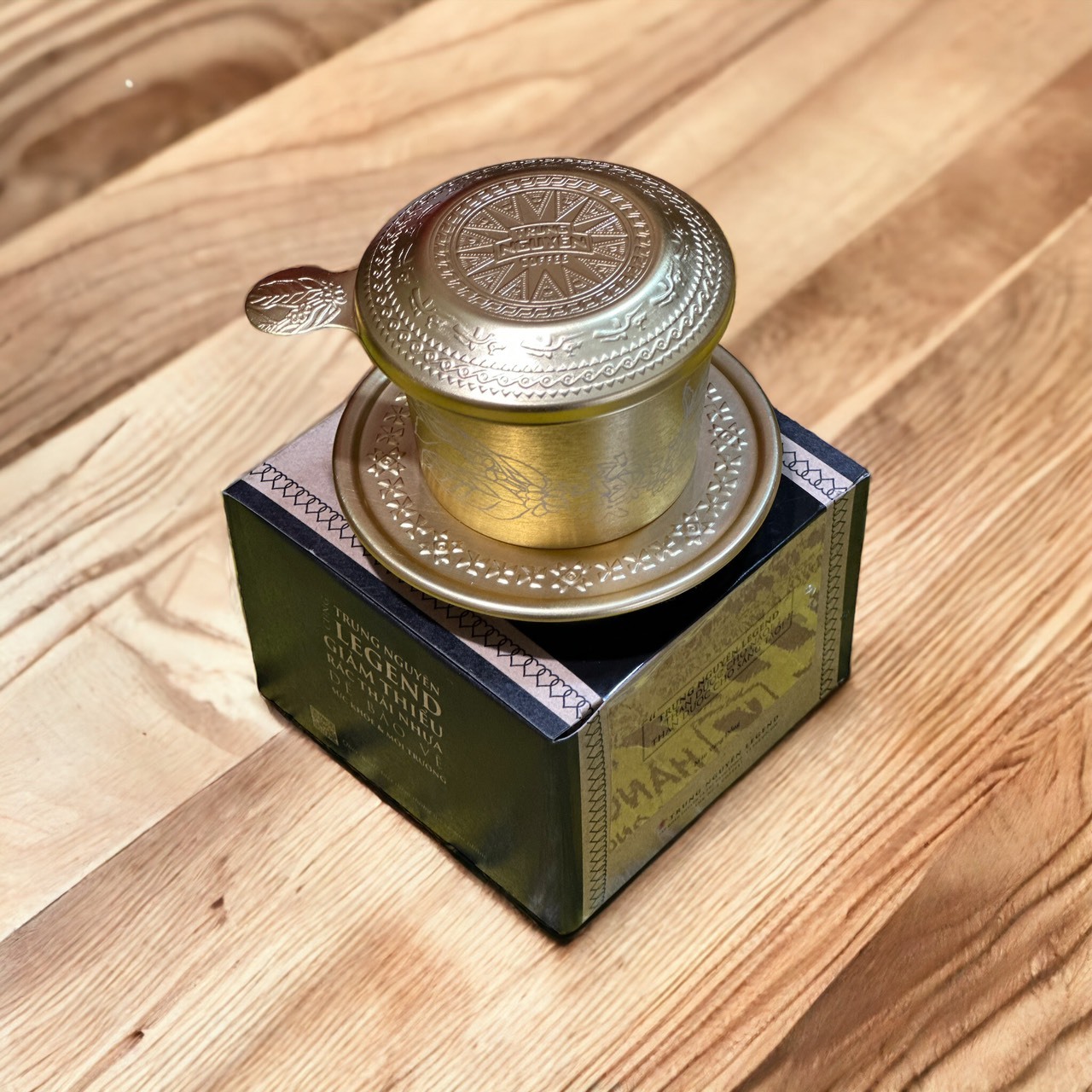 Bộ dụng cụ pha cà phê bằng kim loại đặt trên hộp giấy, nền gỗ sáng màu.