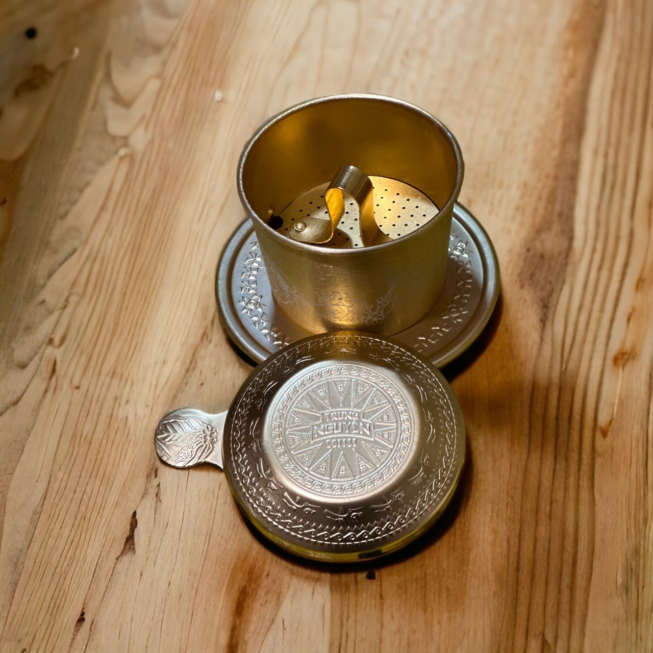 Bộ phin cà phê bằng kim loại trên bàn gỗ, nắp đậy bên cạnh.