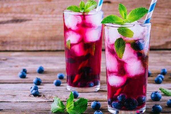 Hai ly nước &eacute;p việt quất, đ&aacute; lạnh, l&aacute; bạc h&agrave; trang tr&iacute;, nền gỗ, quả việt quất rải r&aacute;c xung quanh.