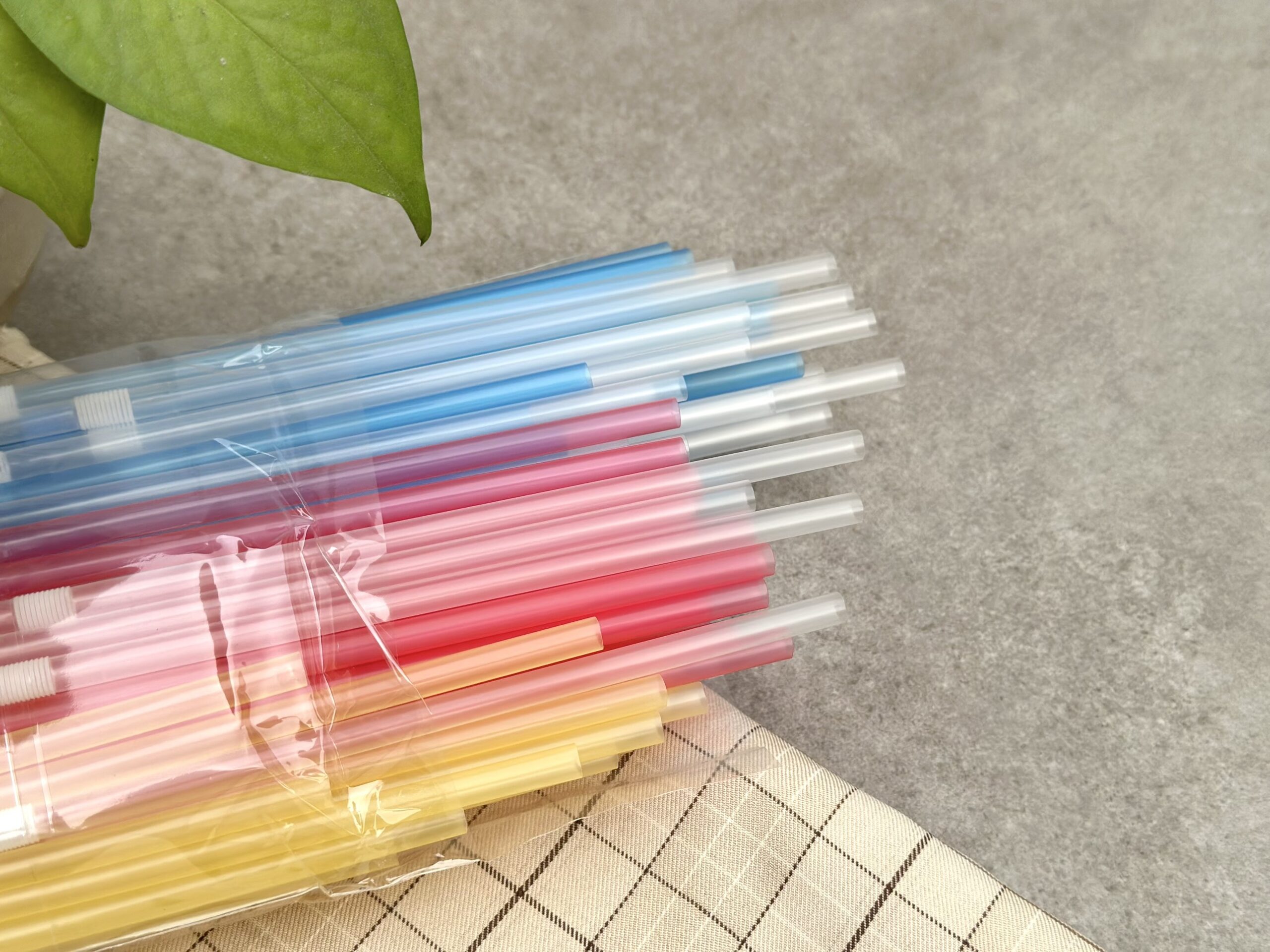 Ống hút nhựa nhiều màu sắc được bọc trong túi nhựa, đặt trên khăn có họa tiết caro và nền xám.
