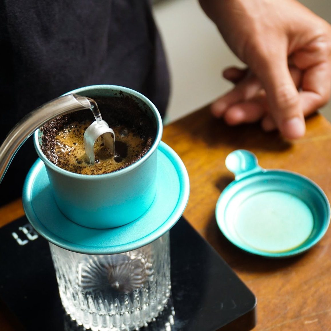 Người pha cà phê phin đang rót nước từ ấm vào phin màu xanh trên ly thủy tinh, bên cạnh là nắp phin.