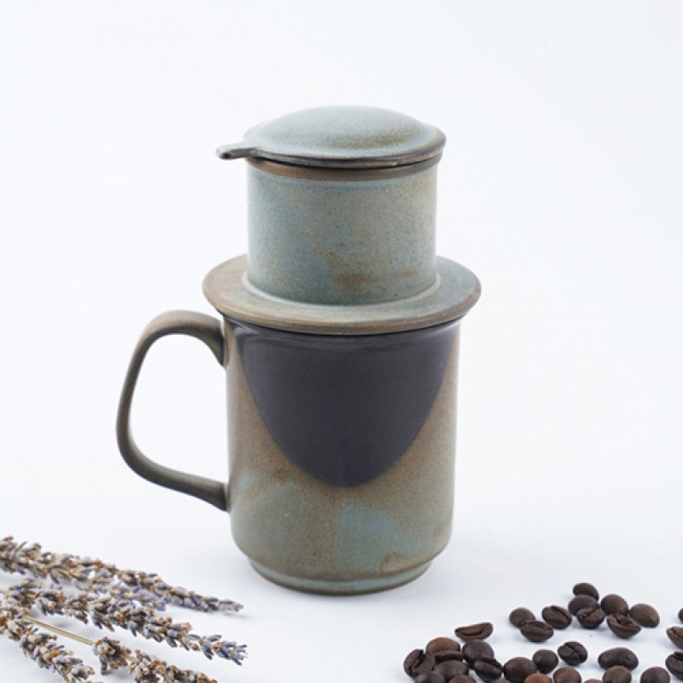 Bộ phin pha cà phê bằng gốm màu xanh, bên cạnh là hạt cà phê và nhánh hoa khô trên nền trắng.