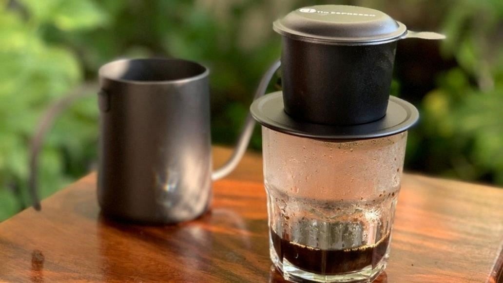 C&agrave; ph&ecirc; phin đang nhỏ giọt v&agrave;o ly thủy tinh tr&ecirc;n b&agrave;n gỗ, b&ecirc;n cạnh l&agrave; một b&igrave;nh đựng kim loại.