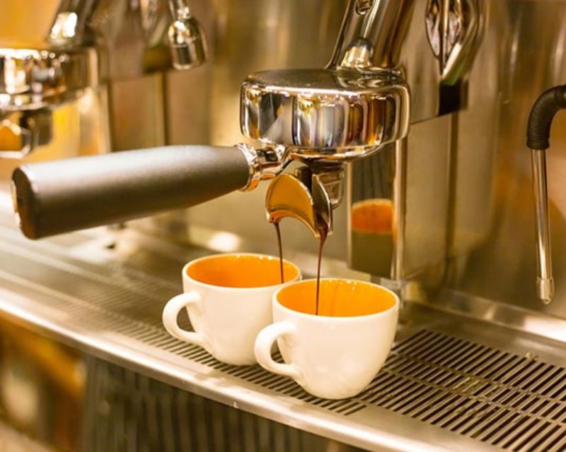 Tuyệt Chiêu Pha Chế Cà Phê Robusta Rang Mộc Đậm Đà Khó Cưỡng 7 Máy pha cà phê đang rót cà phê vào hai tách sứ màu trắng trên khay kim loại.