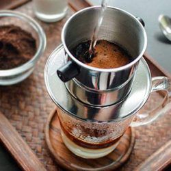 Khám Phá Gu Cà Phê Nguyên Chất - Xu Hướng Thưởng Thức Hiện Đại Của Giới Trẻ! 5 Cà phê phin đang được pha, nước nóng chảy qua phin inox, phía dưới là cốc thủy tinh trên khay gỗ.