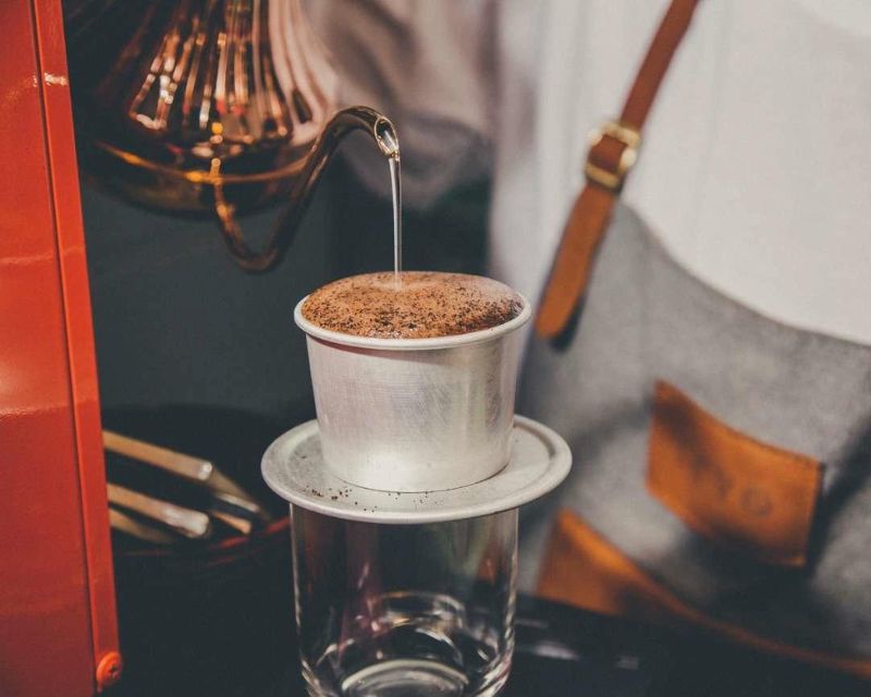 Nước đang được rót từ ấm pha cà phê vào phin trên ly thủy tinh.