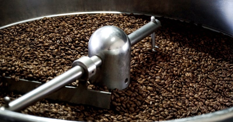 Hạt c&agrave; ph&ecirc; đang được rang trong m&aacute;y c&ocirc;ng nghiệp với c&aacute;nh khuấy kim loại.