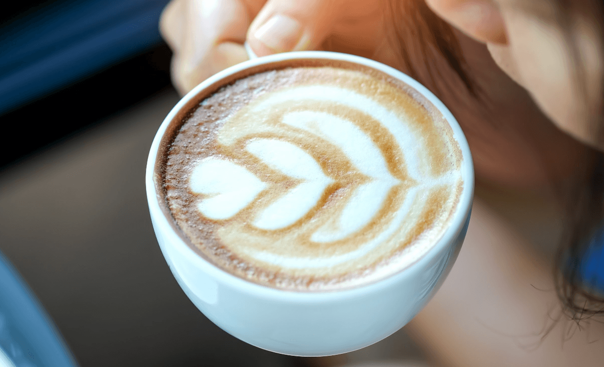 T&aacute;ch c&agrave; ph&ecirc; với lớp bọt sữa được tạo h&igrave;nh nghệ thuật, cầm tr&ecirc;n tay, nền mờ nhạt.