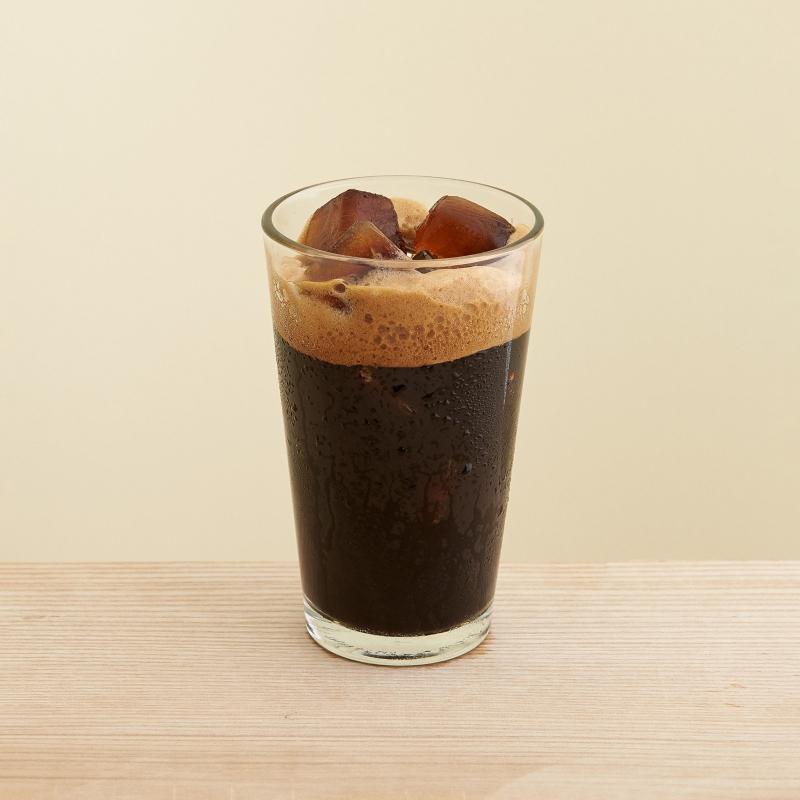 Ly cà phê đá với bọt nổi trên bề mặt, được đặt trên bàn gỗ sáng màu.