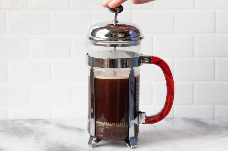 Một c&aacute;i b&igrave;nh pha c&agrave; ph&ecirc; kiểu French press với tay cầm m&agrave;u đỏ, đang được sử dụng tr&ecirc;n mặt b&agrave;n đ&aacute;.