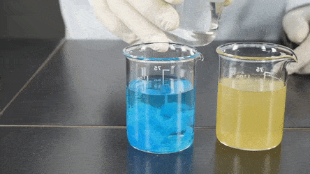 Người đeo găng tay đổ chất lỏng từ cốc thủy tinh vào dung dịch xanh trong cốc, tạo ra sự thay đổi màu sắc.