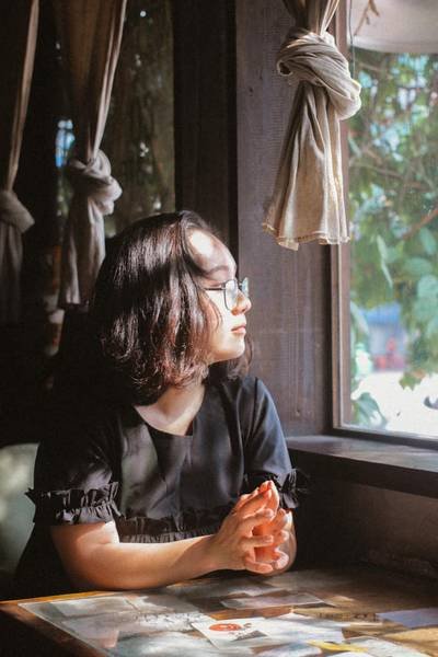 Người ngồi b&ecirc;n cửa sổ, &aacute;nh s&aacute;ng chiếu v&agrave;o, nh&igrave;n ra ngo&agrave;i. Kh&ocirc;ng gian ấm &aacute;p với r&egrave;m cửa v&agrave; c&acirc;y xanh.