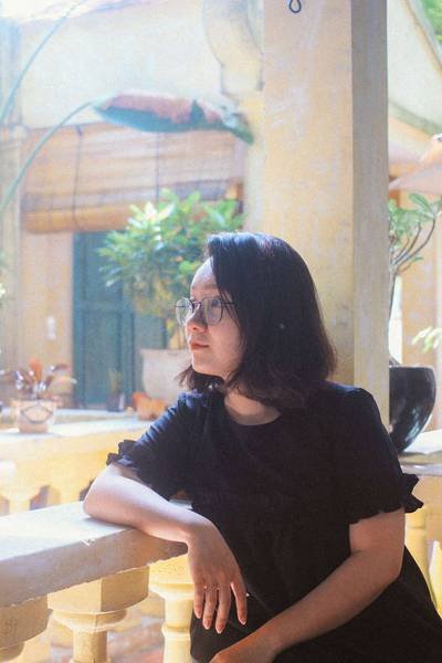 Người phụ nữ t&oacute;c ngắn ngồi tựa v&agrave;o lan can, nh&igrave;n xa xăm, nền l&agrave; c&acirc;y xanh v&agrave; t&ograve;a nh&agrave; cổ k&iacute;nh.