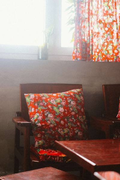 Ghế gỗ với gối tựa hoa văn đỏ tươi, đặt gần cửa sổ c&oacute; r&egrave;m hoa, &aacute;nh s&aacute;ng tự nhi&ecirc;n chiếu v&agrave;o.