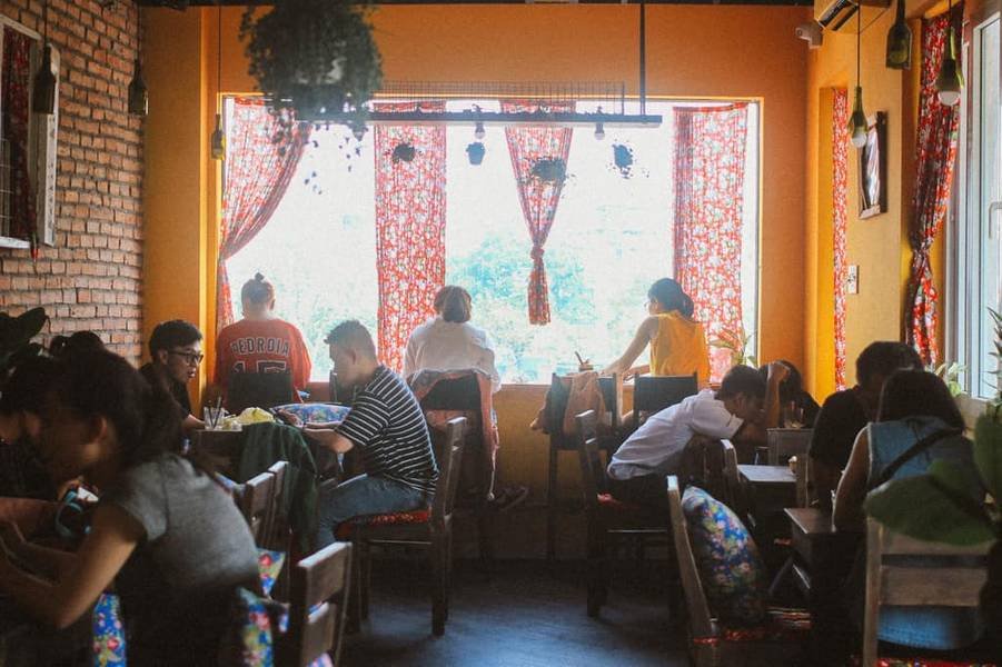 Qu&aacute;n c&agrave; ph&ecirc; với nhiều người ngồi tr&ograve; chuyện v&agrave; l&agrave;m việc, &aacute;nh s&aacute;ng tự nhi&ecirc;n chiếu qua cửa sổ lớn c&oacute; r&egrave;m hoa.