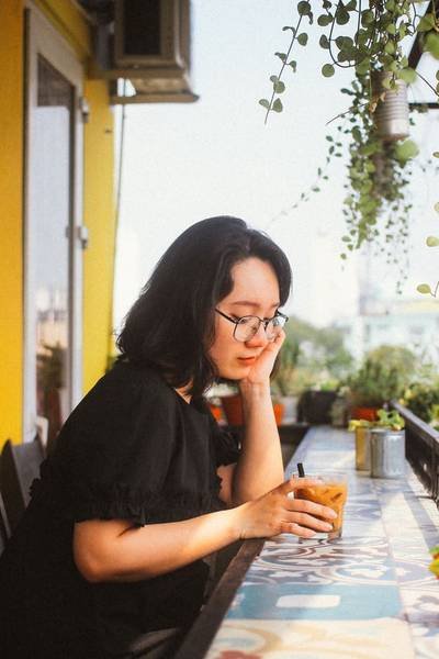 Người phụ nữ ngồi ngo&agrave;i ban c&ocirc;ng, tay cầm ly c&agrave; ph&ecirc;, xung quanh c&oacute; nhiều c&acirc;y xanh v&agrave; hoa trang tr&iacute;.