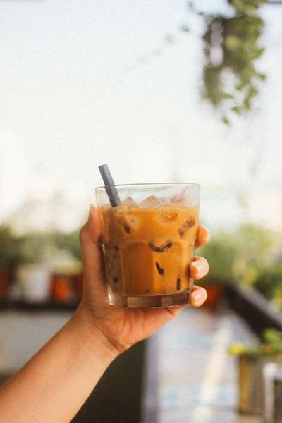 Tay cầm ly c&agrave; ph&ecirc; sữa đ&aacute; với ống h&uacute;t, nền l&agrave; cảnh vật mờ ảo v&agrave; c&acirc;y xanh.